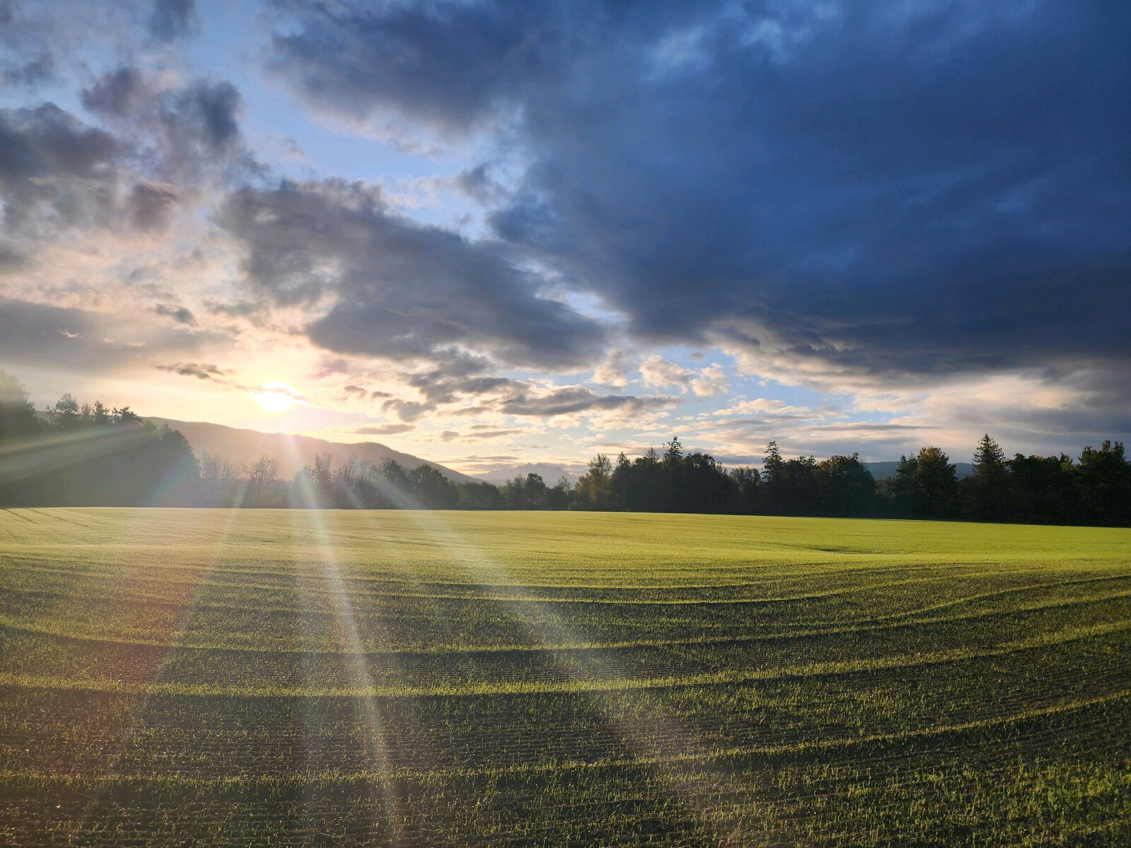 Sunlight breaking through clouds over a green field bordered by trees and hills